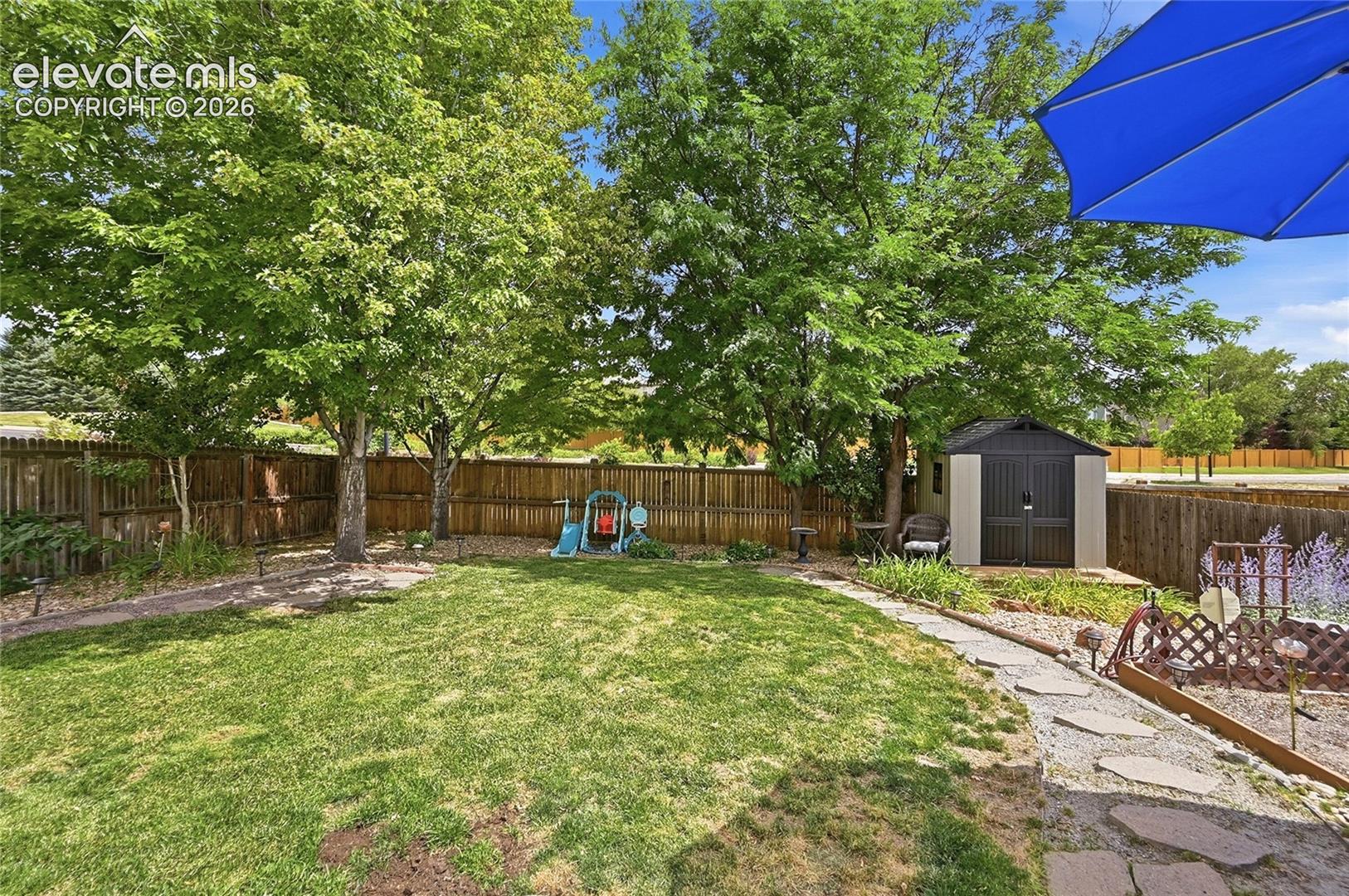 Fenced backyard featuring a manicured lawn, stone paver pathway, mature trees, and a detached storage shed