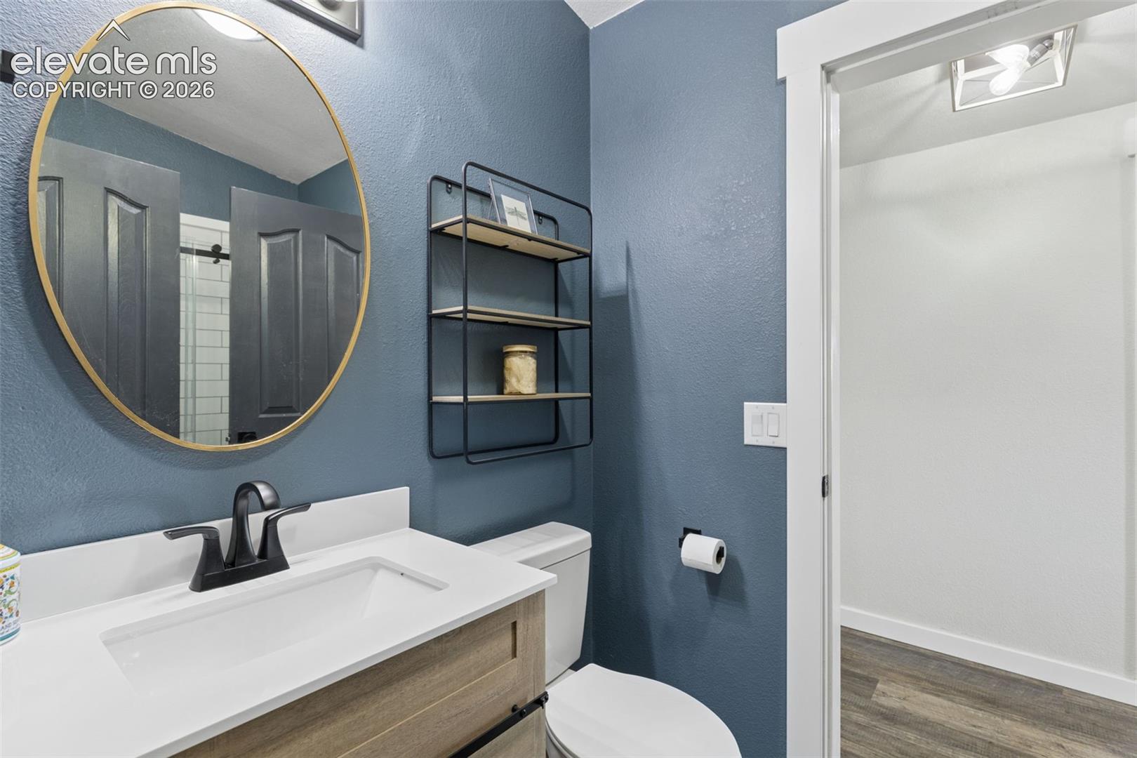 Vanity featuring a light wood-finish cabinet, rectangular basin, and stone-composite countertop paired with a matte black faucet, oval gold-rimmed mirror, and matte black wall-mounted shelving unit