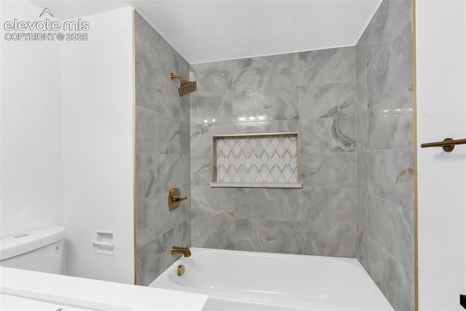 Bathtub surround featuring large-format stone-patterned tile, integrated recessed shelving with decorative geometric inlay, and brass-tone fixtures