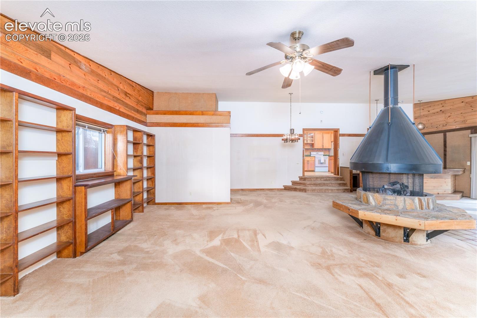 Spacious room featuring a central fireplace with a stone hearth, extensive built-in shelving, wood-paneled accents, and a ceiling fan