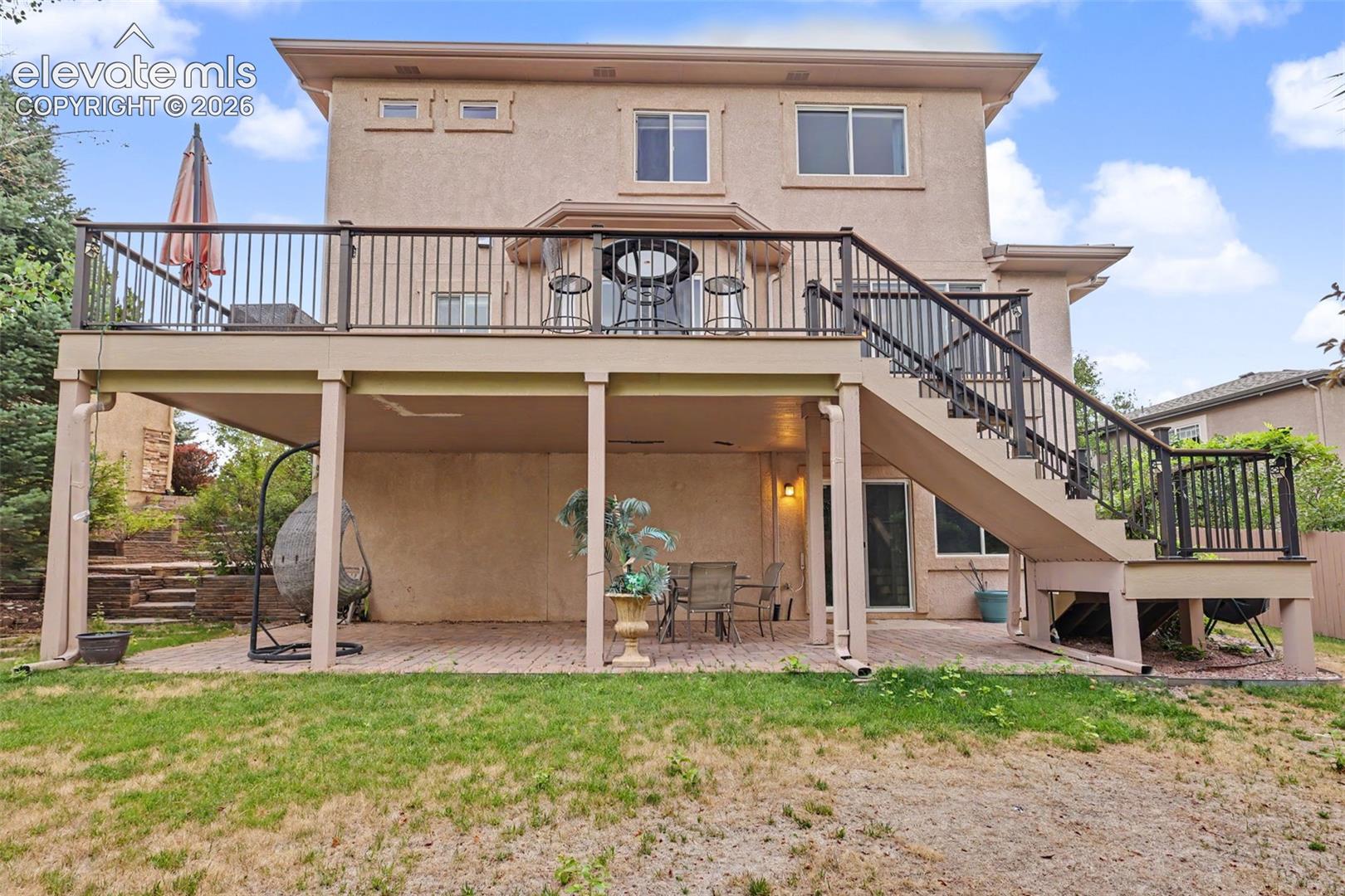Two-story stucco exterior featuring an elevated deck with black railings and stairs, providing access to a ground-level patio with paver flooring