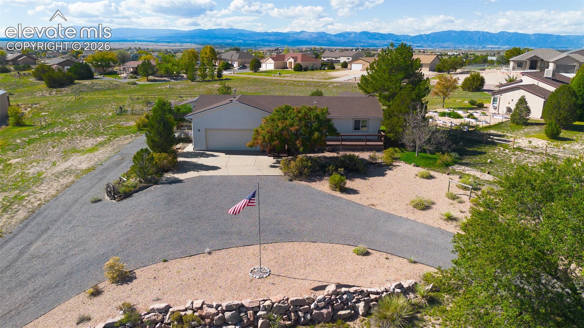 Single-story residence featuring a two-car garage, gravel driveway, stone retaining wall, and mountain range backdrop