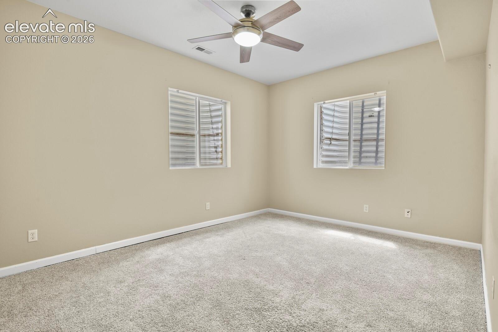 Spacious room featuring neutral carpeting, light-toned wall paint, two windows with horizontal blinds, and a ceiling fan with light fixture