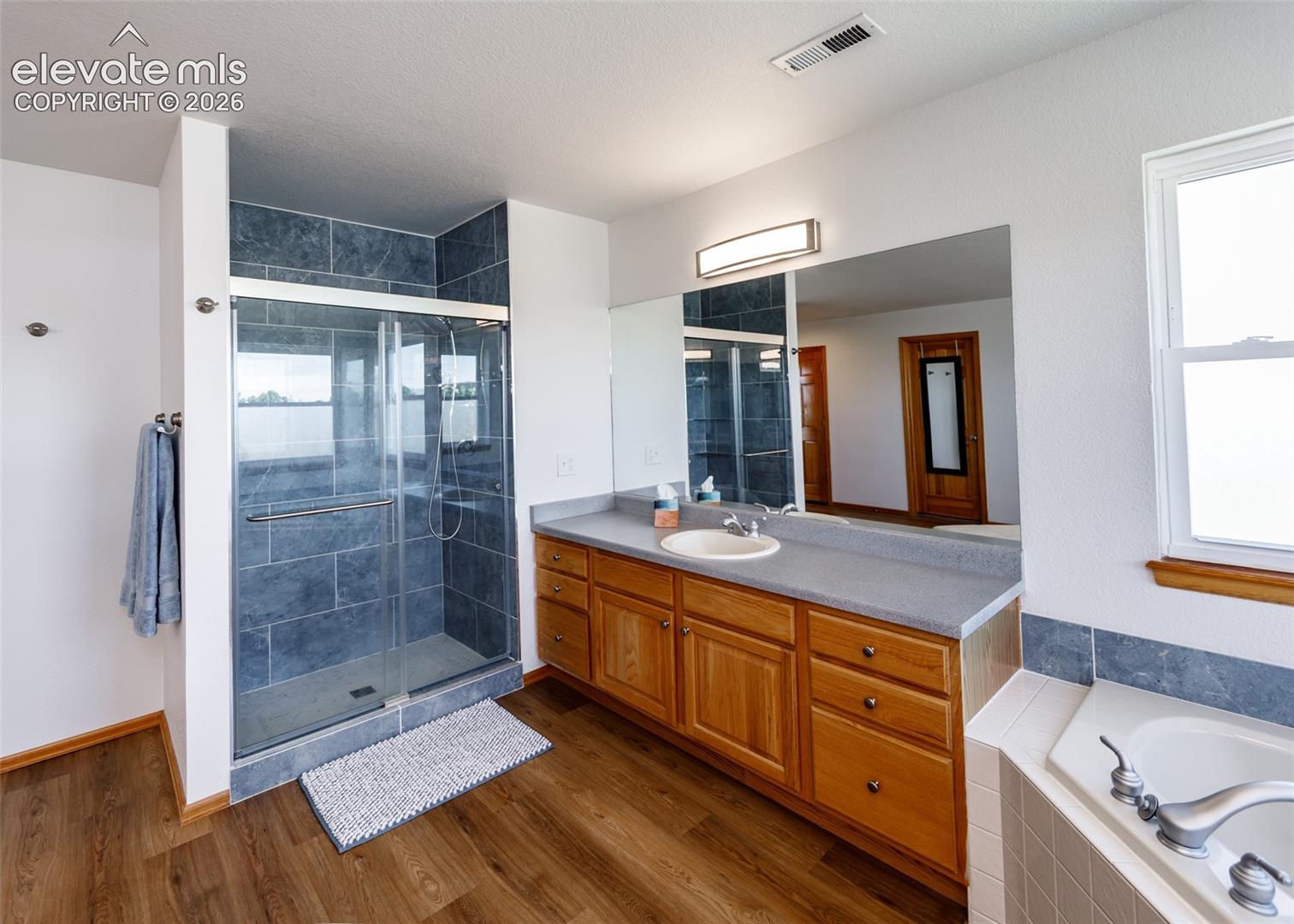 Glass-enclosed shower with dark tile surround, single vanity with wood cabinetry and countertop, large mirror, and wood-finish flooring