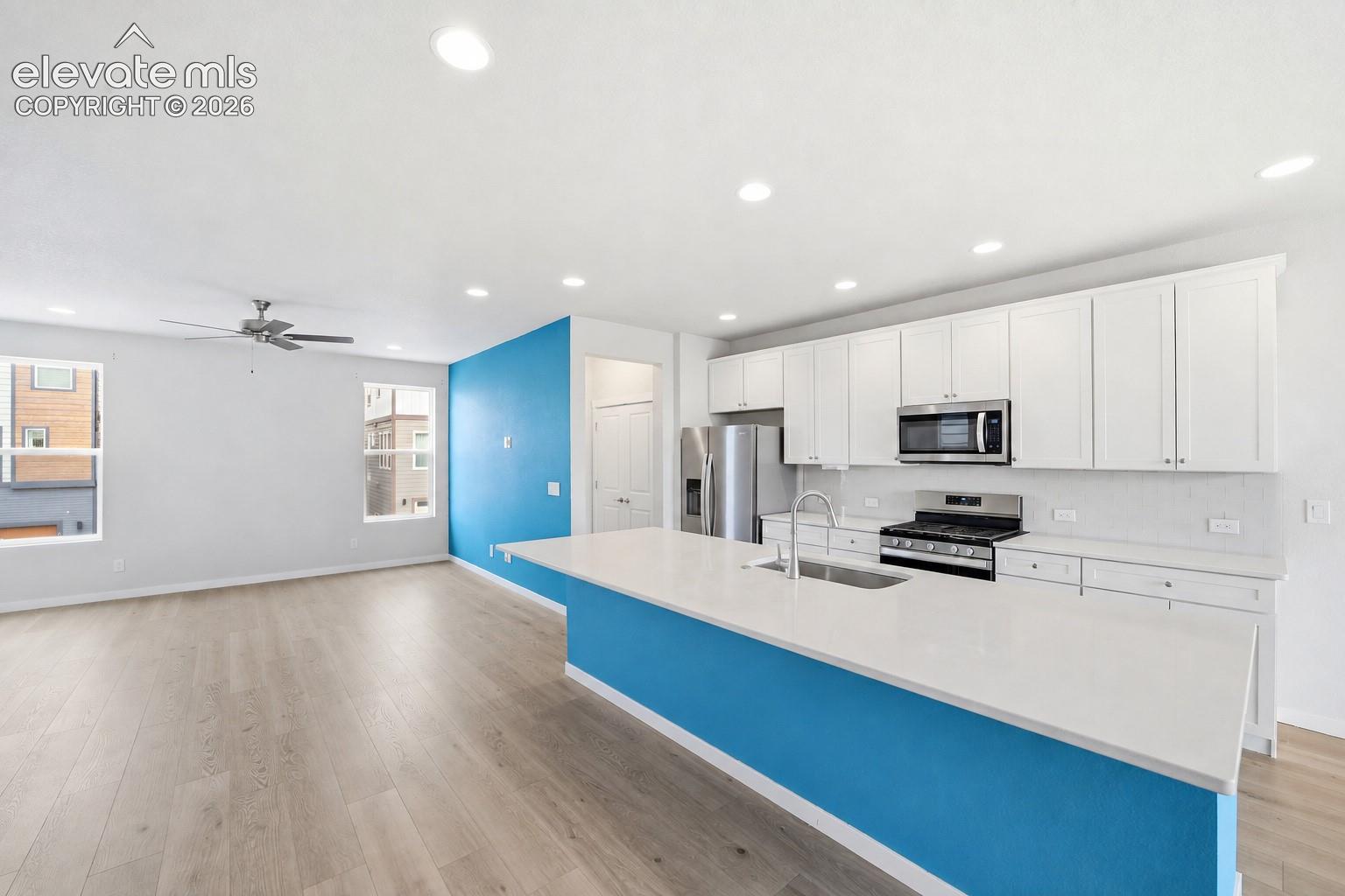 Kitchen island with a white countertop and integrated sink, stainless steel appliances, white cabinetry, wood-finish flooring, and recessed lighting