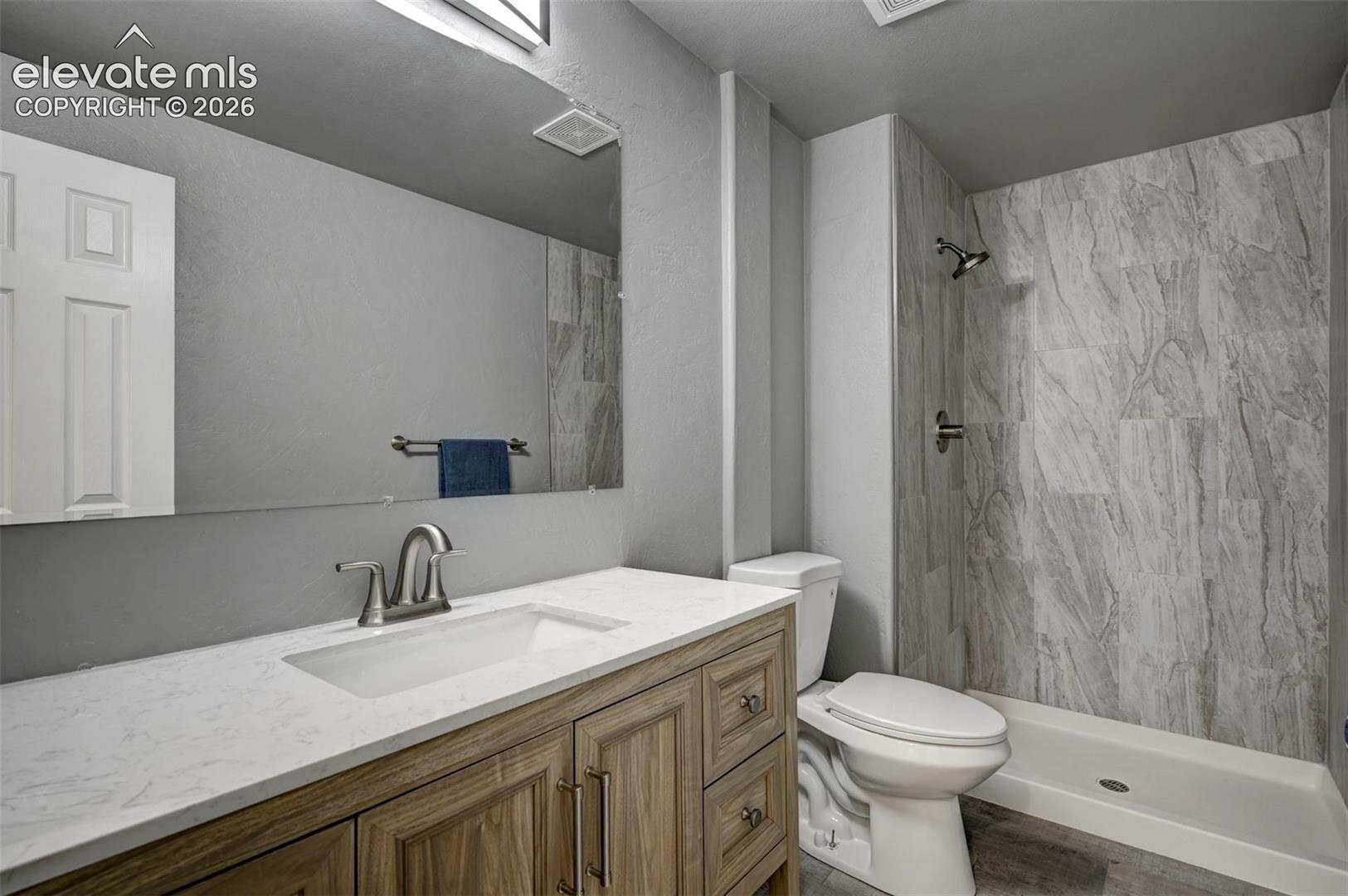 Bathroom featuring a vanity with wood-finish cabinetry, a white countertop with an integrated basin, a large wall-mounted mirror, and a shower enclosure with light-toned wall tile