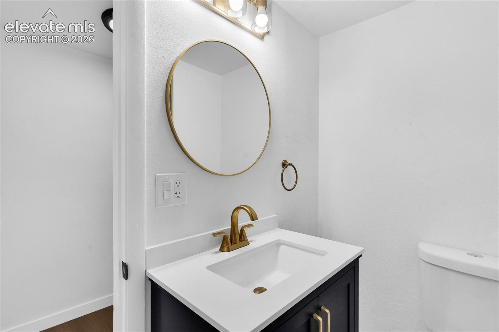 Vanity featuring a stone-composite countertop, undermount sink, brass-finish faucet, circular brass-framed mirror, and dark-toned cabinetry with matching hardware