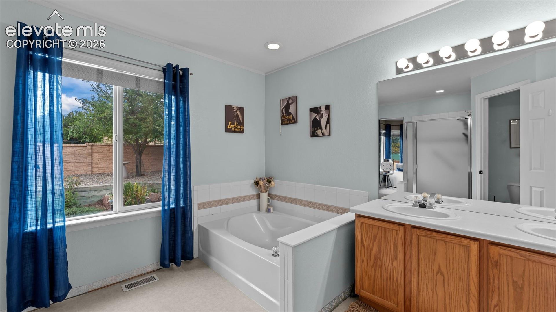 Bathroom featuring a deep-soaking tub with tile surround, double-sink vanity with wood-finish cabinetry, full-width mirror, and multi-bulb wall lighting