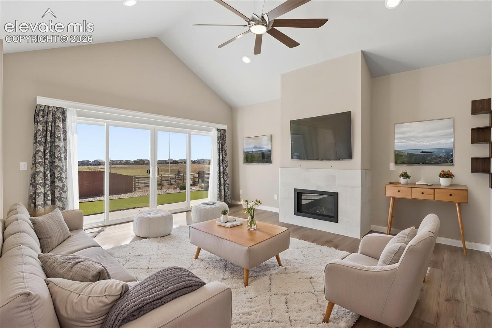 Vaulted ceilings, large sliding glass doors, fireplace with light-toned masonry surround, wood-finish flooring, and a multi-blade ceiling fan