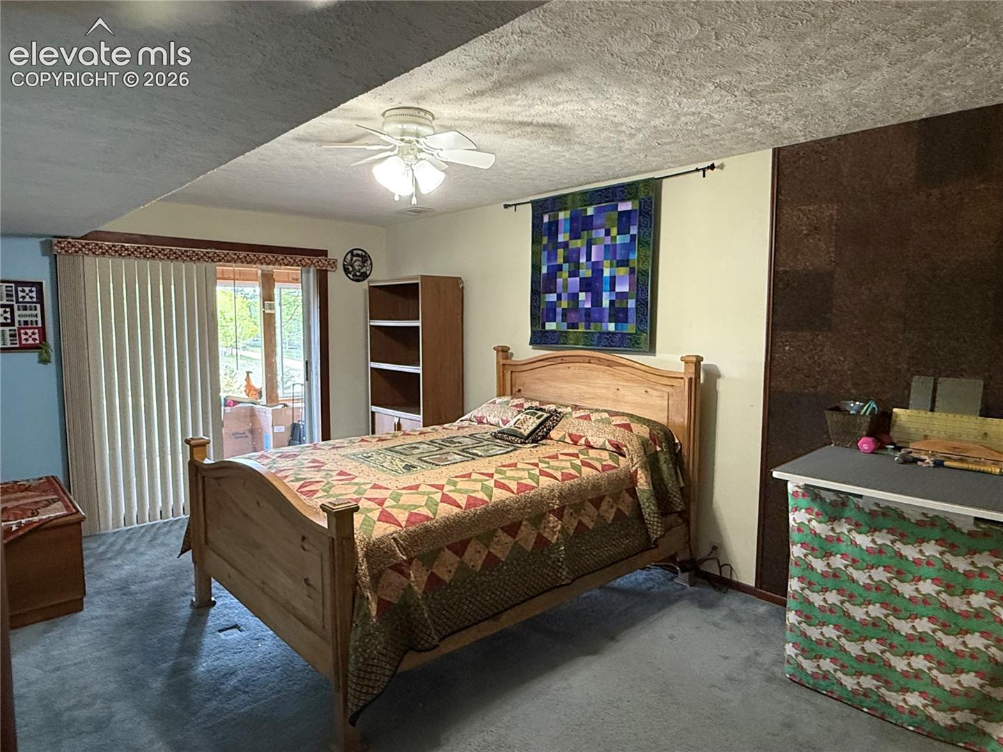 Carpeted room featuring a ceiling fan with integrated lighting, a sliding glass door with vertical blinds, and a built-in shelving unit
