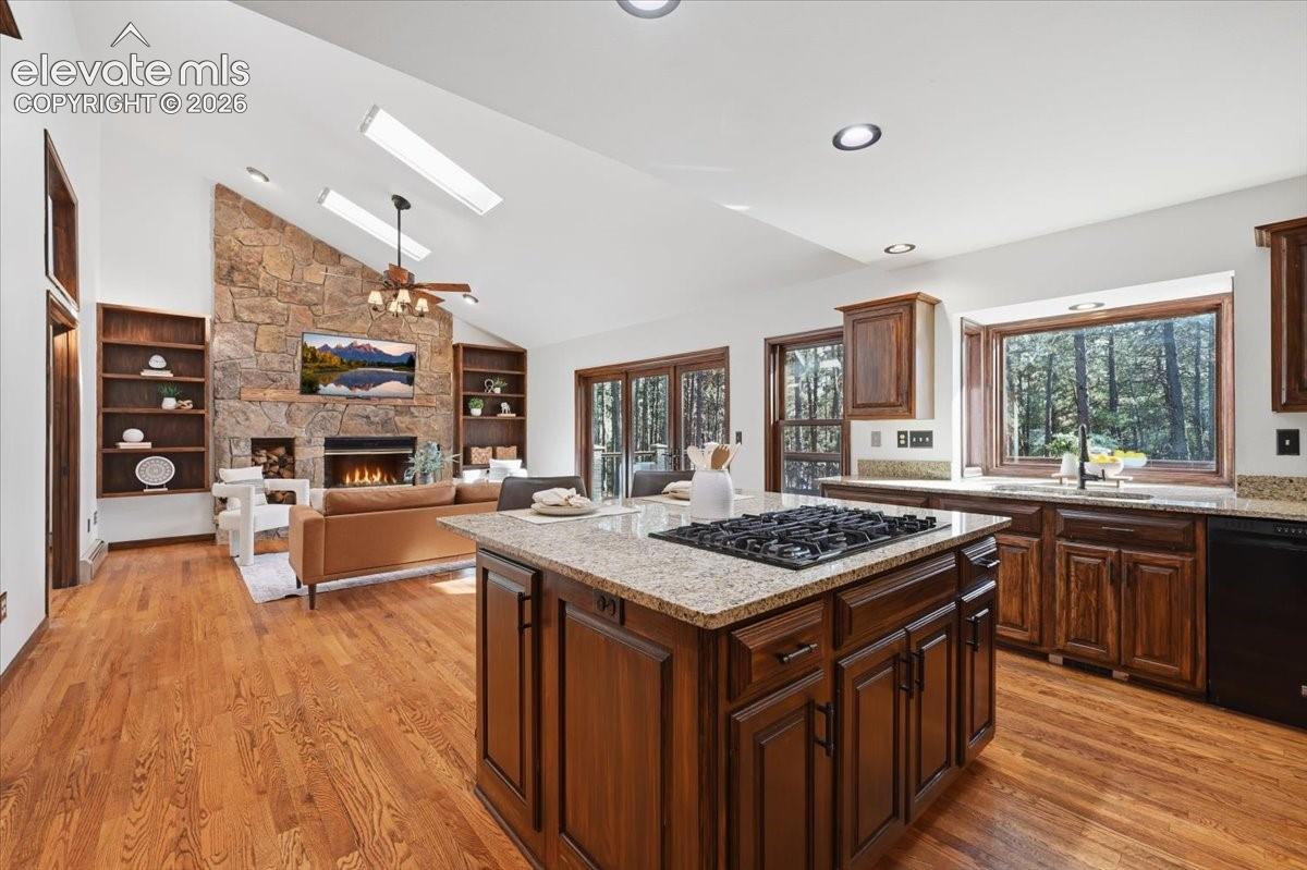 Vaulted ceiling with skylights, stone-faced fireplace, built-in shelving, wood-finish flooring, and a kitchen island with cooktop