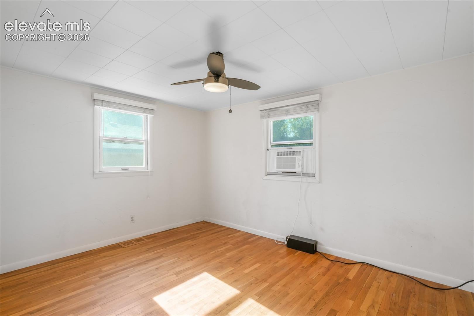 Hardwood-finish flooring, two windows with blinds, ceiling fan with integrated lighting, and a wall-mounted air conditioning unit