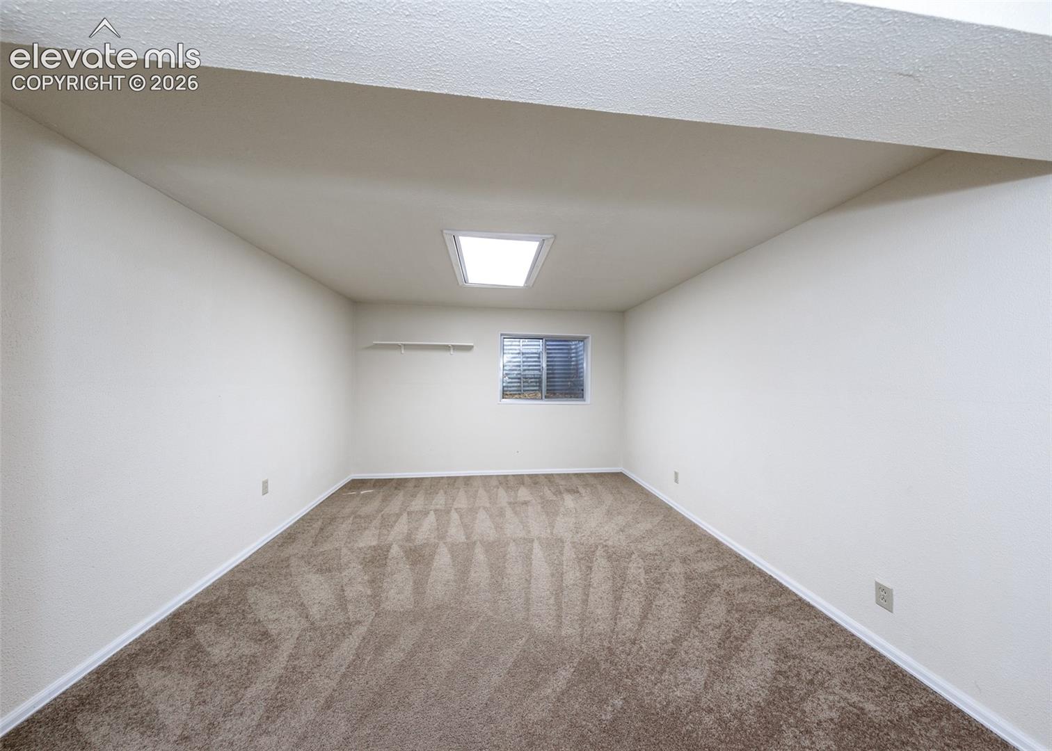 Room interior featuring neutral wall-to-wall carpeting, a vaulted ceiling, a centered skylight, and a built-in wall shelf