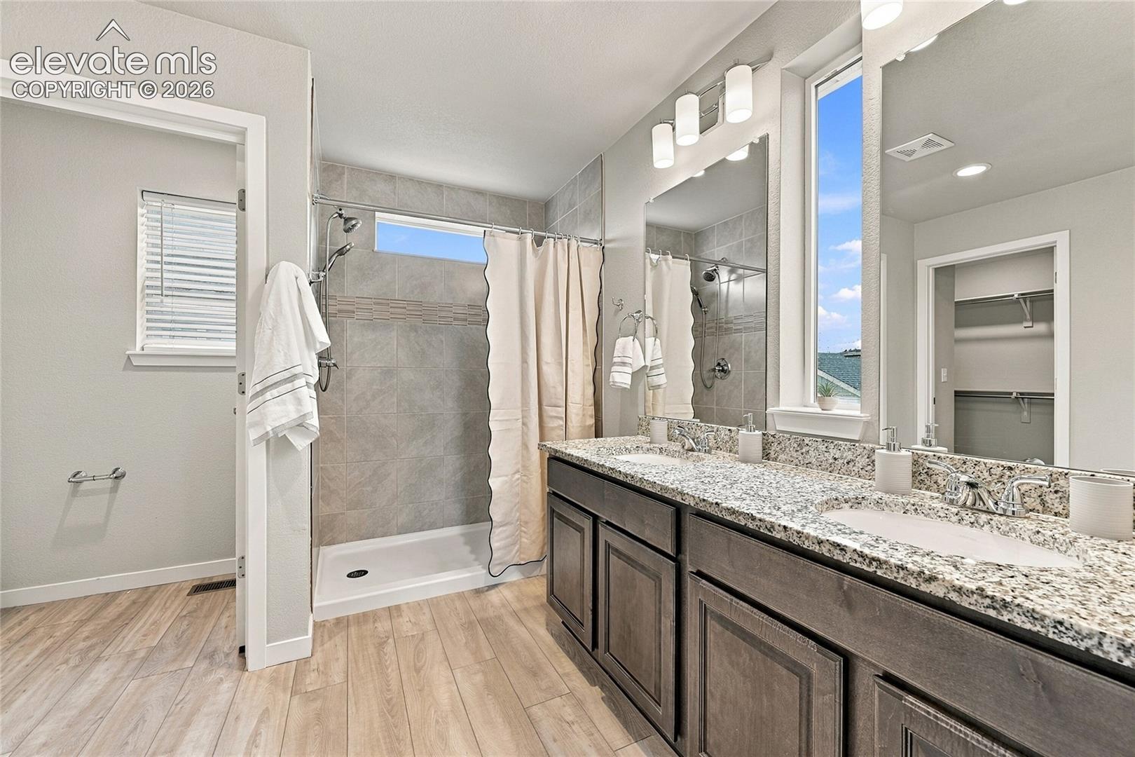 Dual vanities with stone-finish countertops, wood-finish flooring, tiled shower with transom window, and ample natural light