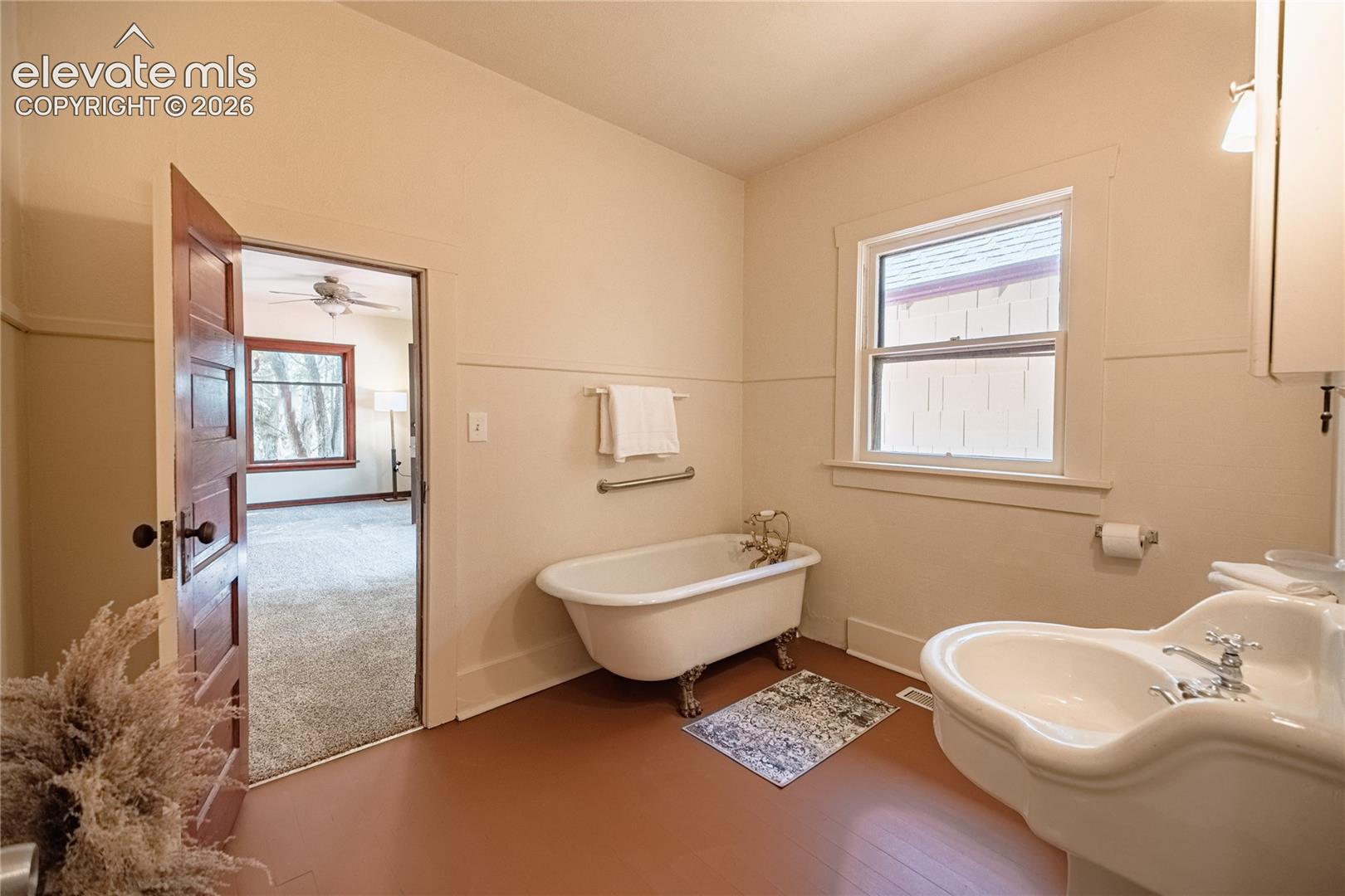 Clawfoot bathtub with brass fixtures, pedestal sink with chrome faucets, window with white trim, and wood-finish flooring