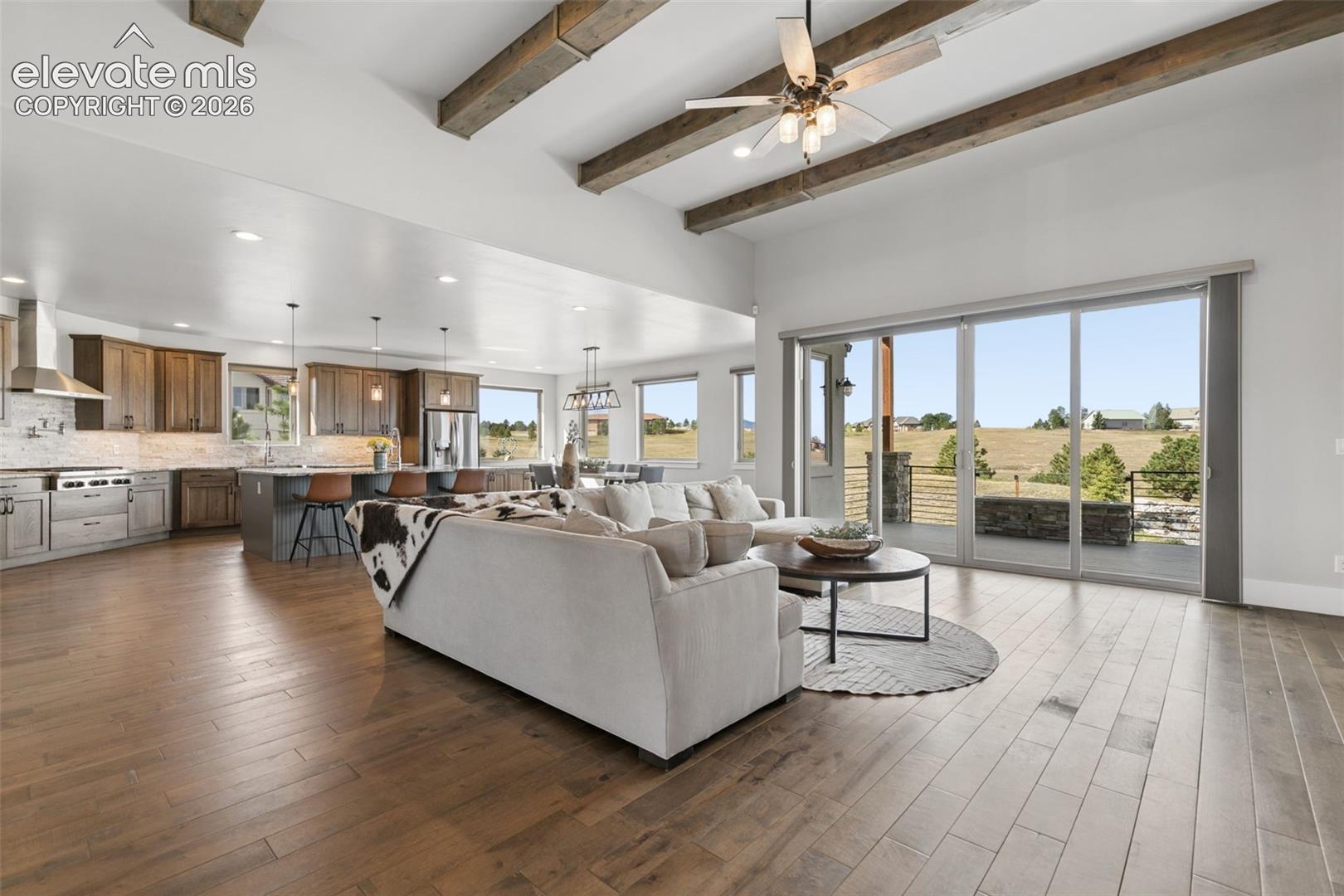 Vaulted ceilings with exposed wood beams, expansive wood-finish flooring, a kitchen featuring an island with seating, stainless steel appliances, and large sliding glass doors opening to the exterior