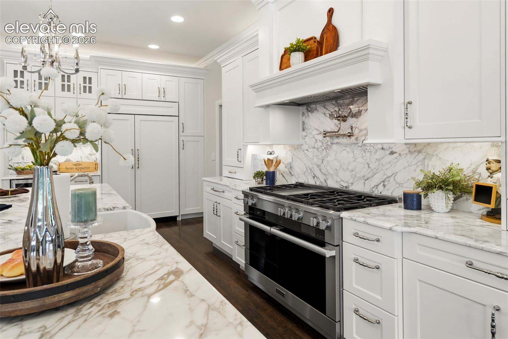 Custom cabinetry with crown molding, natural stone countertops, professional-grade gas range, stone tile backsplash with pot filler, and wood-finish flooring