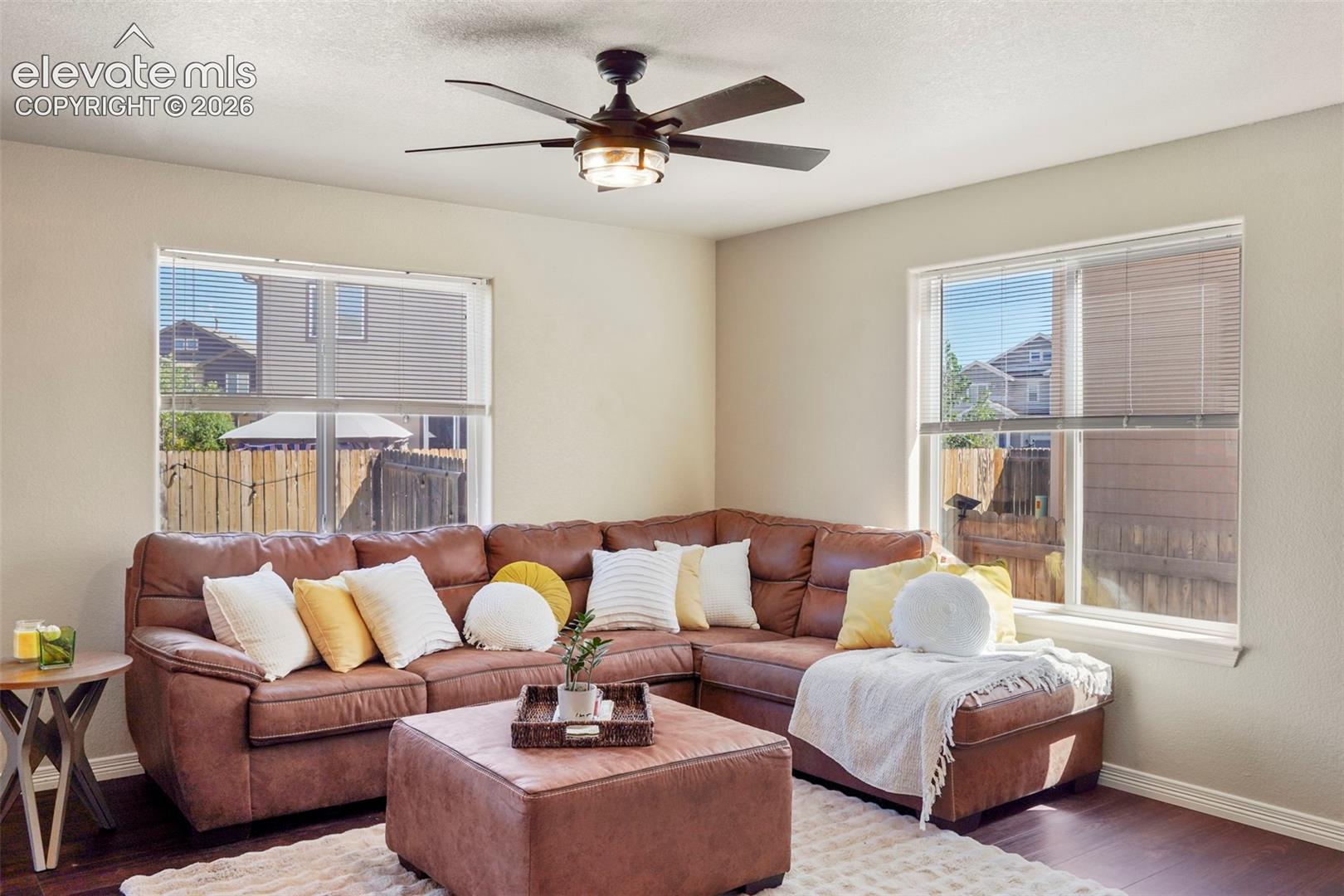 Bright living area featuring wood-finish flooring, two large windows with blinds, a ceiling fan with integrated lighting, and neutral wall tones