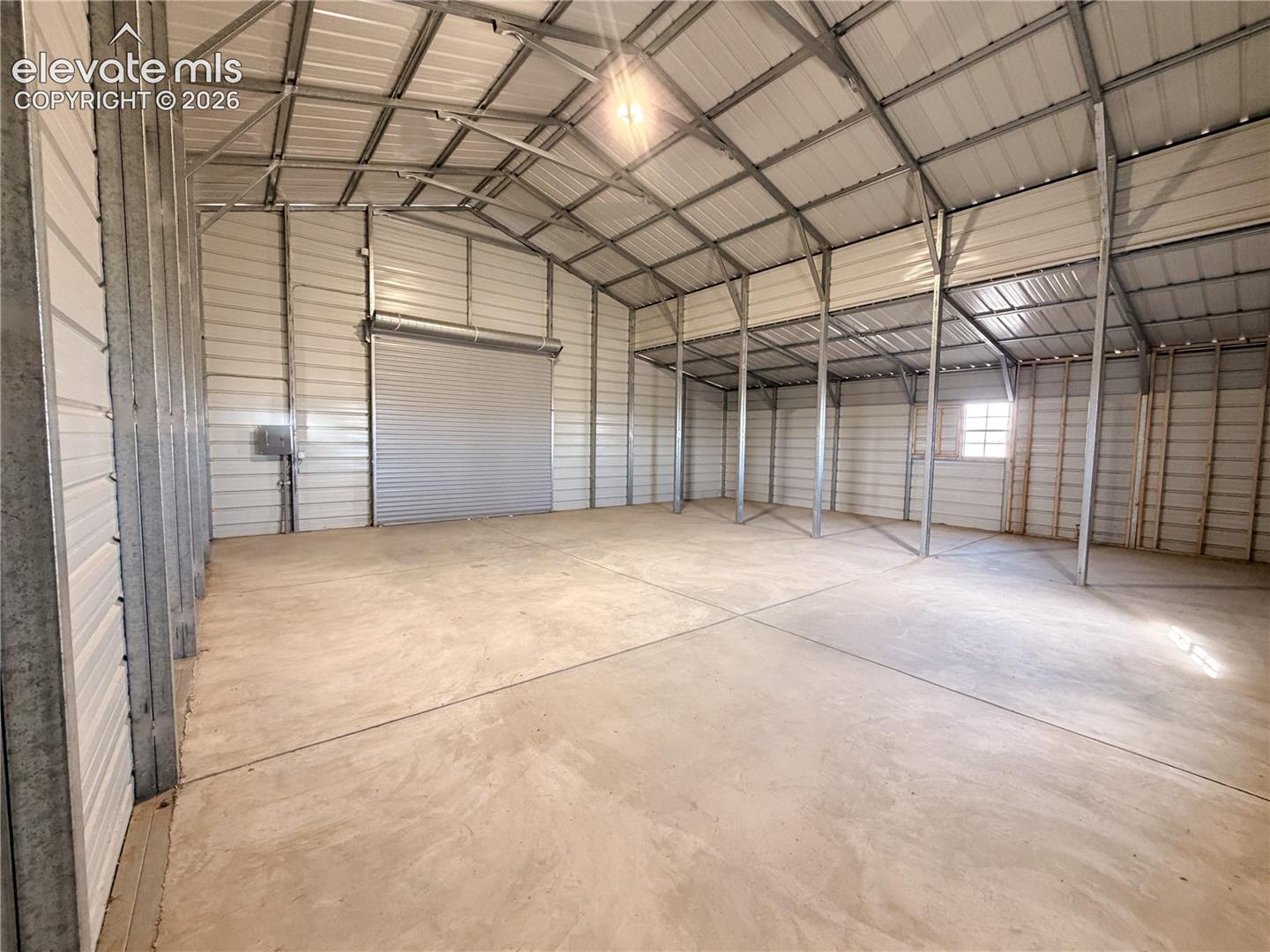 Spacious metal-frame structure featuring concrete flooring, high vaulted ceilings, a roll-up garage door, and metal wall paneling