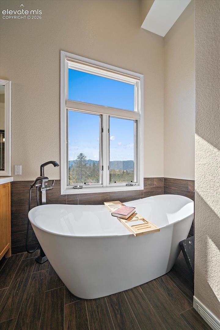 Freestanding white soaking tub with a floor-mounted matte black faucet, dark wood-finish tile flooring, horizontal dark wood-finish tile wall accent, and tall window with mountain scenery