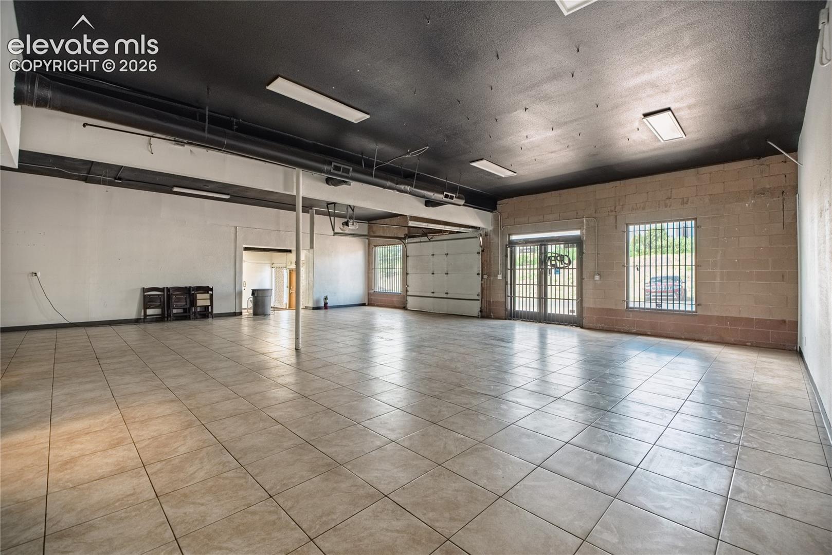 Spacious interior with neutral-toned tile flooring, exposed ductwork, brick accent wall, and a roll-up door