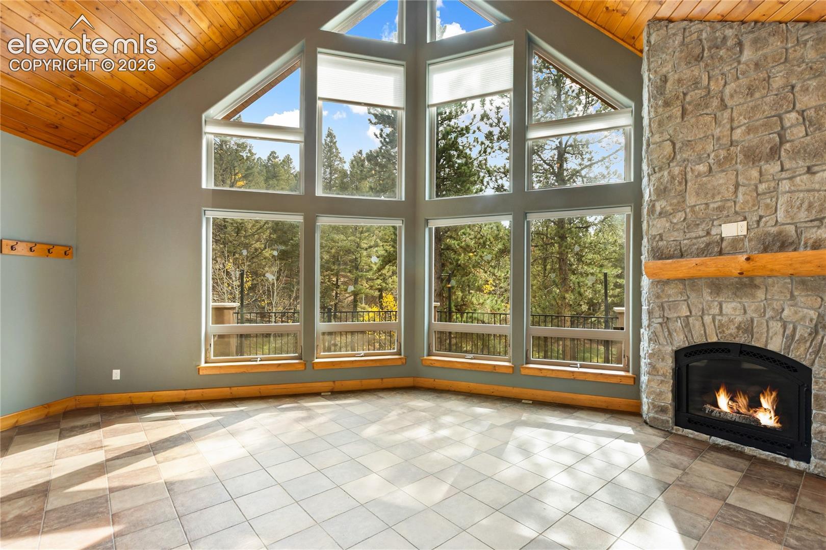 Vaulted wood-paneled ceiling, floor-to-ceiling multi-pane windows, natural stone fireplace, wood mantel, and tile flooring
