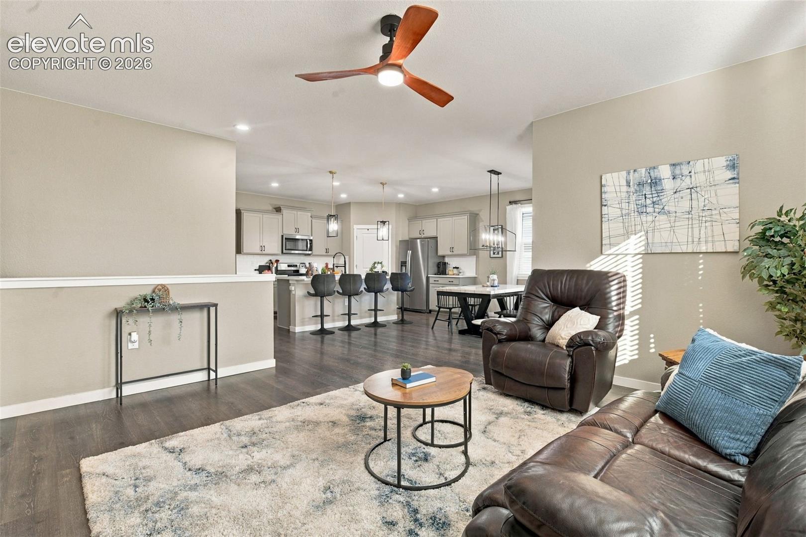 Open-concept interior featuring wood-finish flooring, a three-blade ceiling fan with integrated lighting, shaker-style kitchen cabinetry, a central kitchen island with bar seating, and pendant lighting fixtures