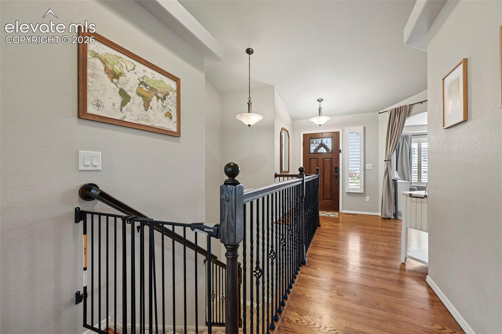 Hardwood flooring, wrought iron baluster railing, wood-finish newel post, pendant lighting, and multi-pane front door with decorative glass inlay