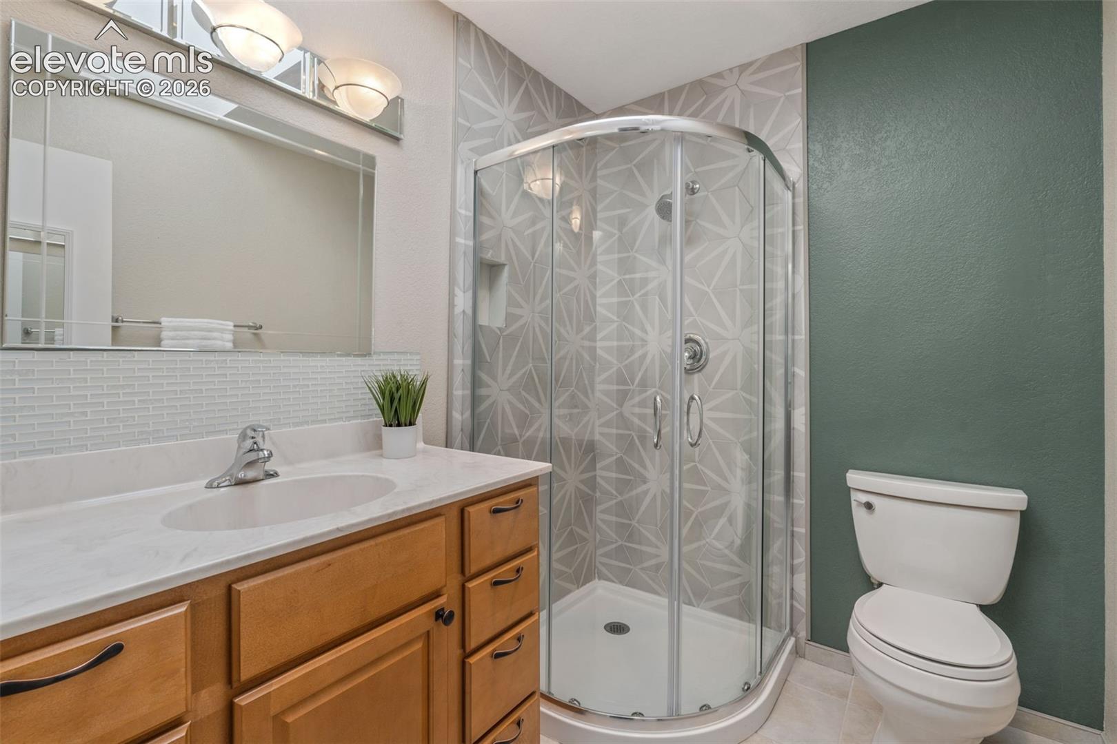 Bathroom featuring a curved glass shower enclosure with geometric-patterned tile surround, light-toned wood vanity, solid surface countertop, integrated sink, and polished chrome fixtures