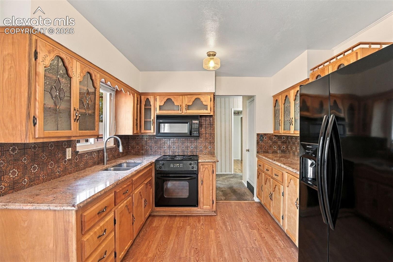 Kitchen featuring natural wood cabinetry with decorative glass inserts, patterned tile backsplash, stone-finish countertops, black built-in appliances, and wood-finish flooring