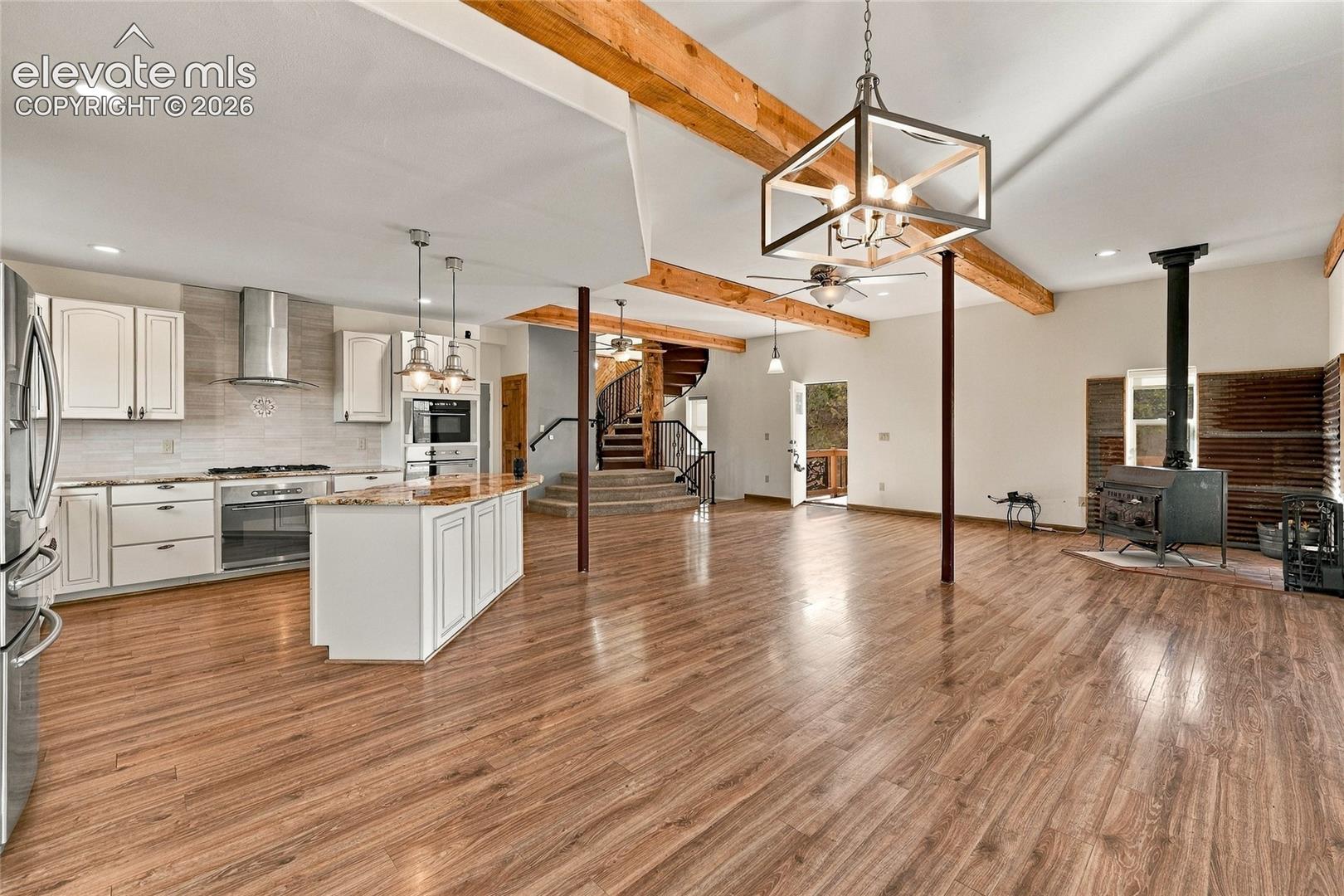 Open-concept interior featuring wood-finish flooring, exposed wooden ceiling beams, a kitchen island with stone countertops, a freestanding wood-burning stove, and a central spiral staircase