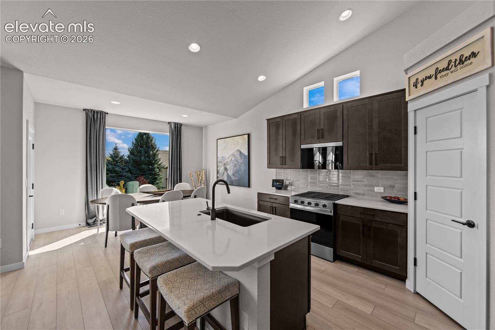Kitchen featuring a central island with a solid-surface countertop and undermount sink, dark-toned cabinetry, stainless steel appliances, neutral backsplash, and vaulted ceilings with recessed lighting