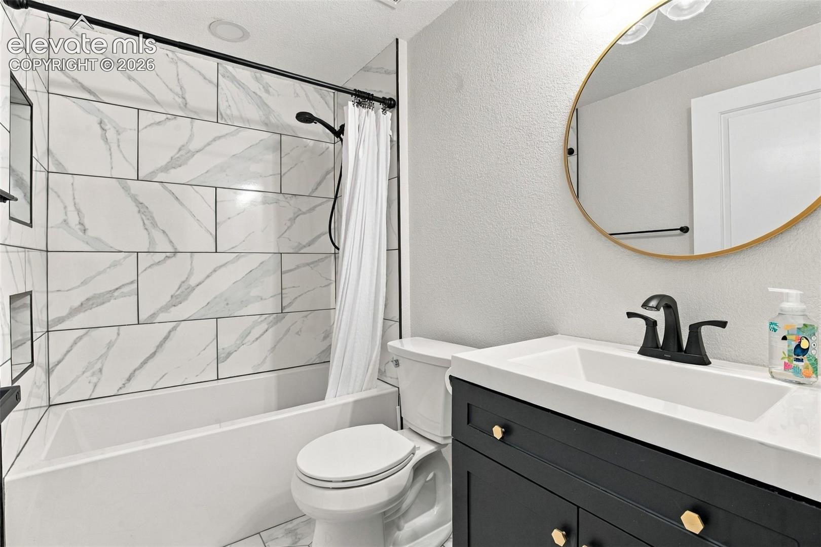 Bathroom featuring a dark vanity with a white countertop, matte black faucet, wall-mounted circular mirror with a brass frame, and bathtub with large-format patterned wall tile and matte black hardware