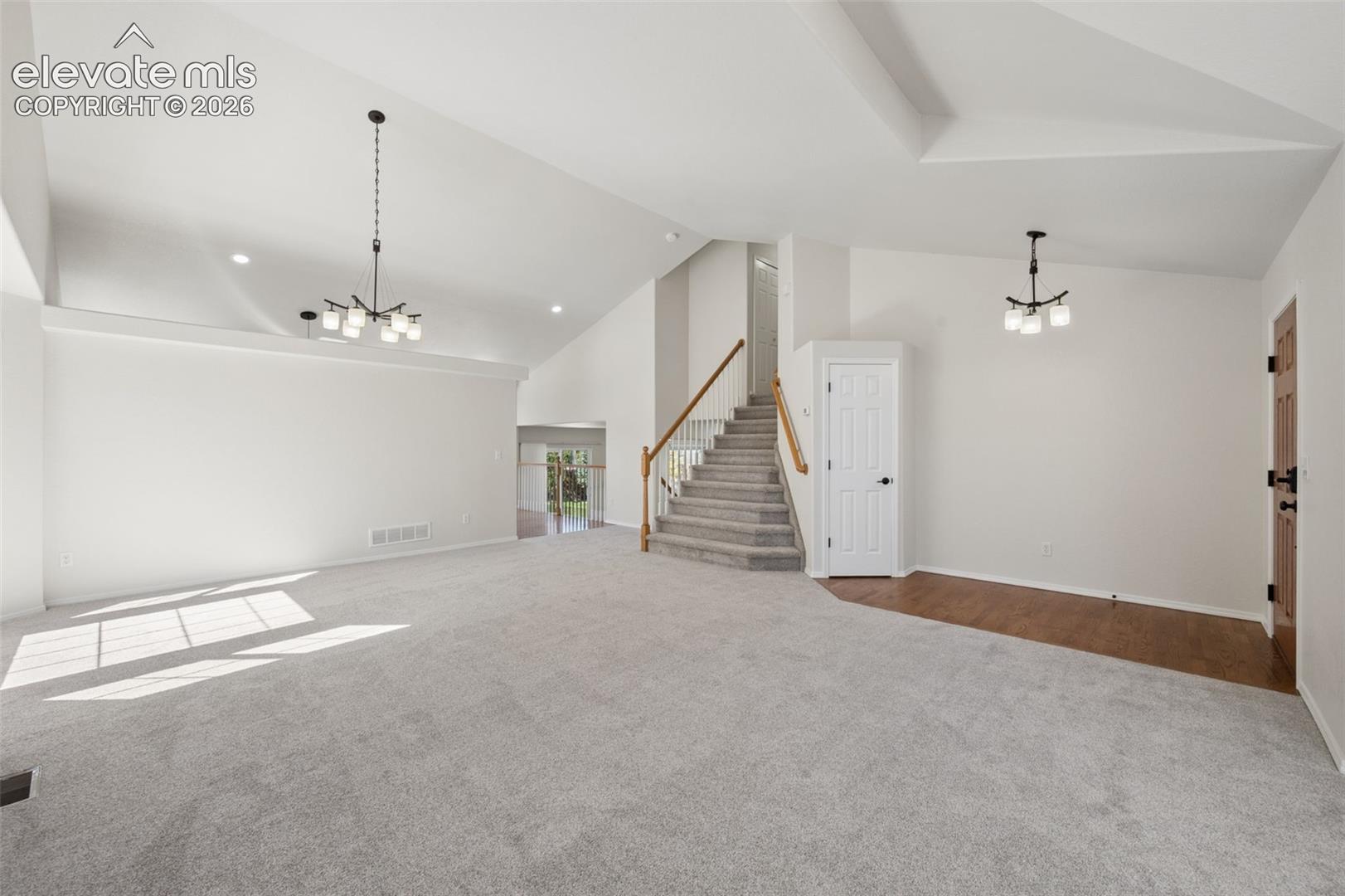 Vaulted ceilings, carpeted flooring, staircase with wood-finish handrail, dual chandelier lighting, and hardwood entryway flooring