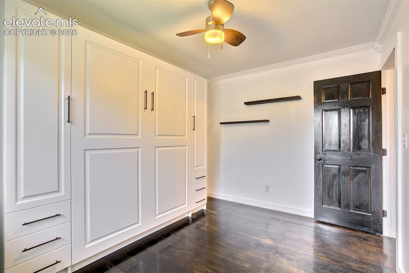 Built-in white cabinetry with recessed-panel doors and matte black hardware, dark wood-finish flooring, two fixed wall-mounted floating shelves, and a ceiling fan with integrated lighting