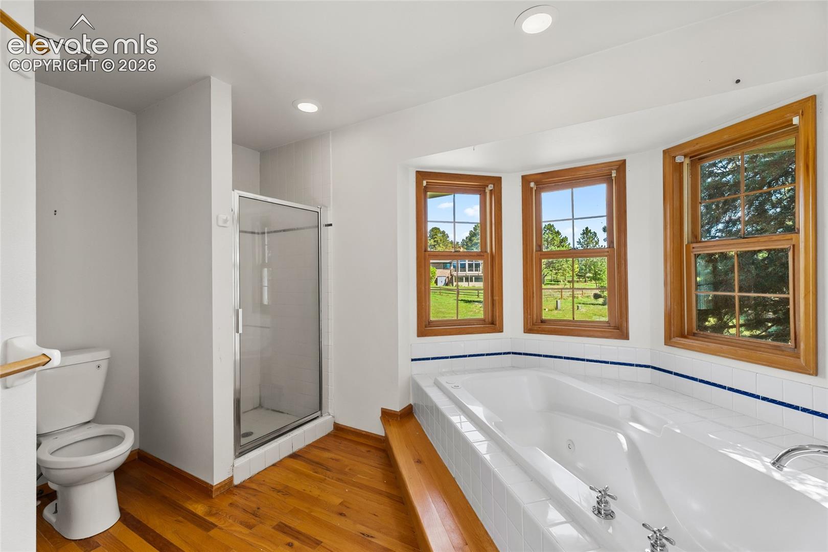 Soaking tub with tile surround, wood-finish flooring, three windows with wood trim, and a glass-enclosed shower