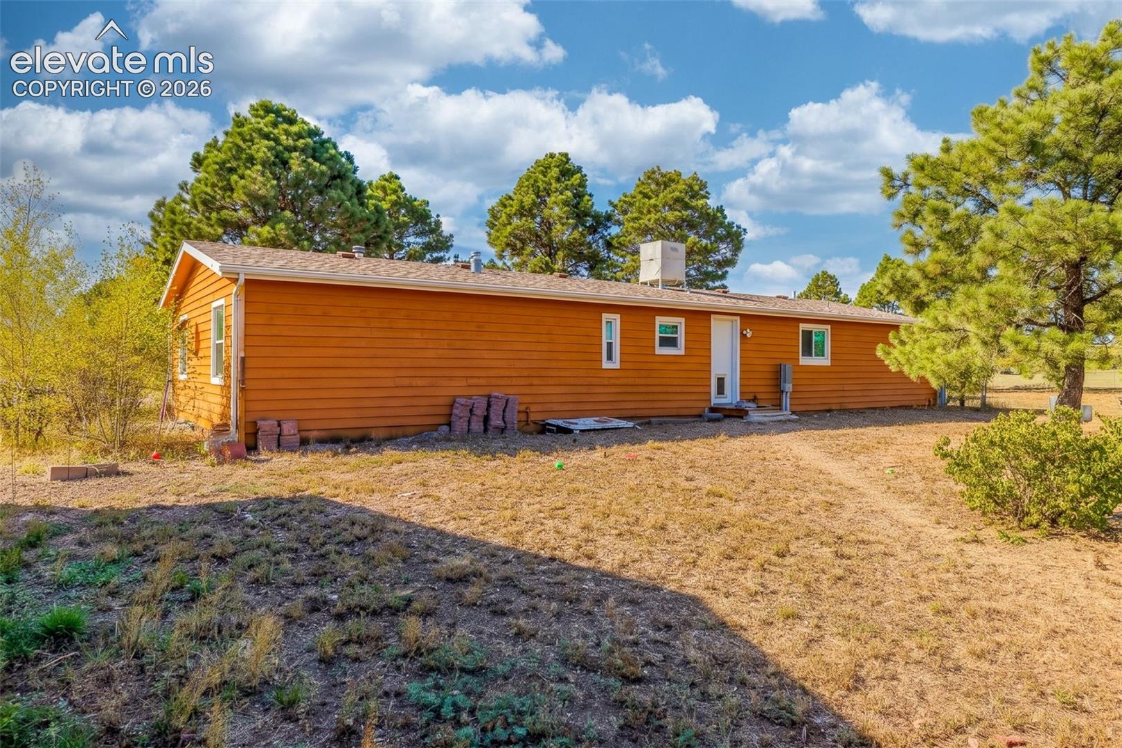 Single-story residence featuring horizontal wood siding, a composite shingle roof, and exterior roof-mounted evaporative cooling unit, situated on a lot with mature pine trees