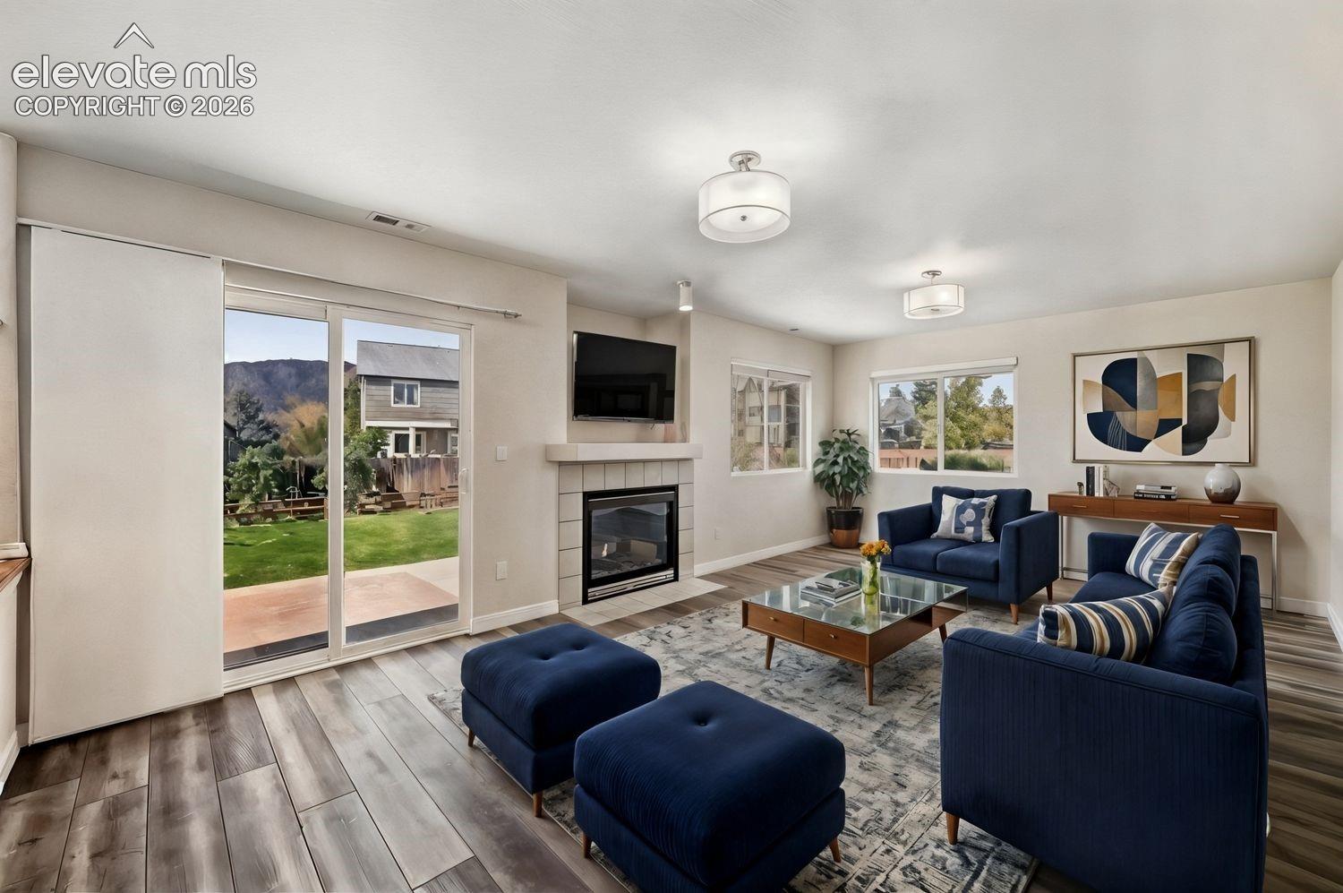 Living area featuring wood-finish flooring, a fireplace with tile surround and a mantel, dual drum-style ceiling light fixtures, and a glass sliding door providing outdoor access