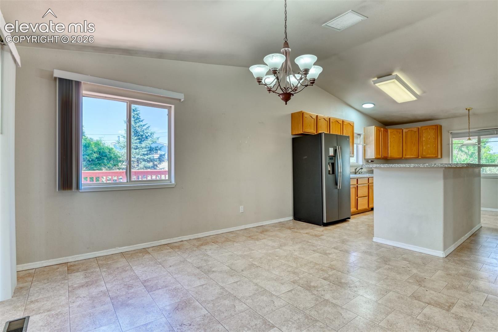 Vaulted ceiling with decorative multi-arm chandelier, neutral tile flooring, kitchen featuring wood cabinetry and stone-surfaced breakfast bar
