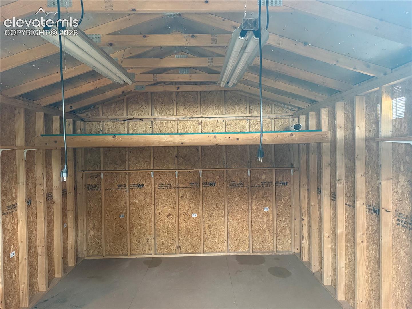 Outbuilding featuring exposed wooden stud framing, a built-in loft storage shelf, and dual suspended fluorescent lighting fixtures