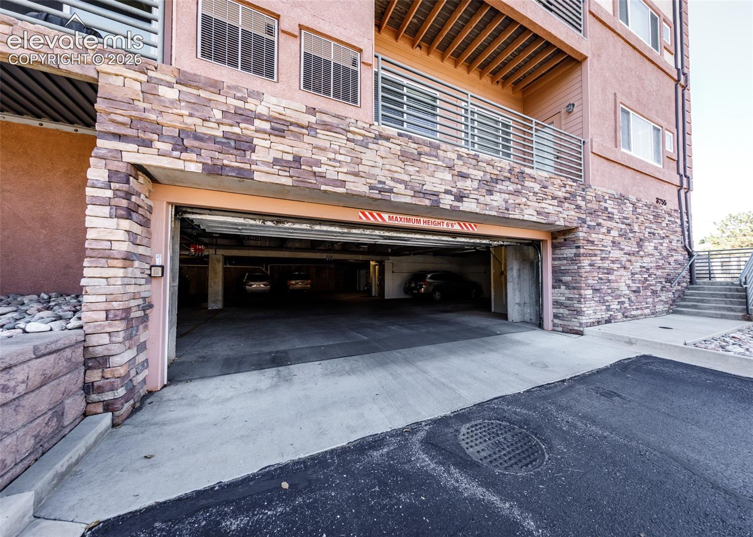 Covered parking structure featuring stacked stone masonry facade, concrete driveway, and upper-level balcony with metal railing