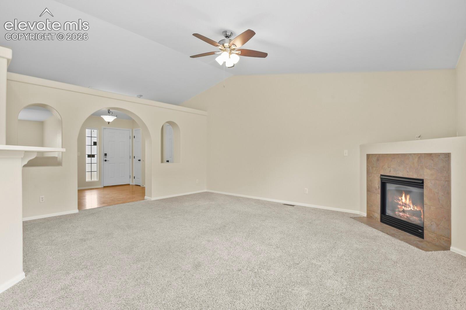 Spacious living area featuring vaulted ceilings, a ceiling fan, plush carpeting, a tiled fireplace, and arched wall details