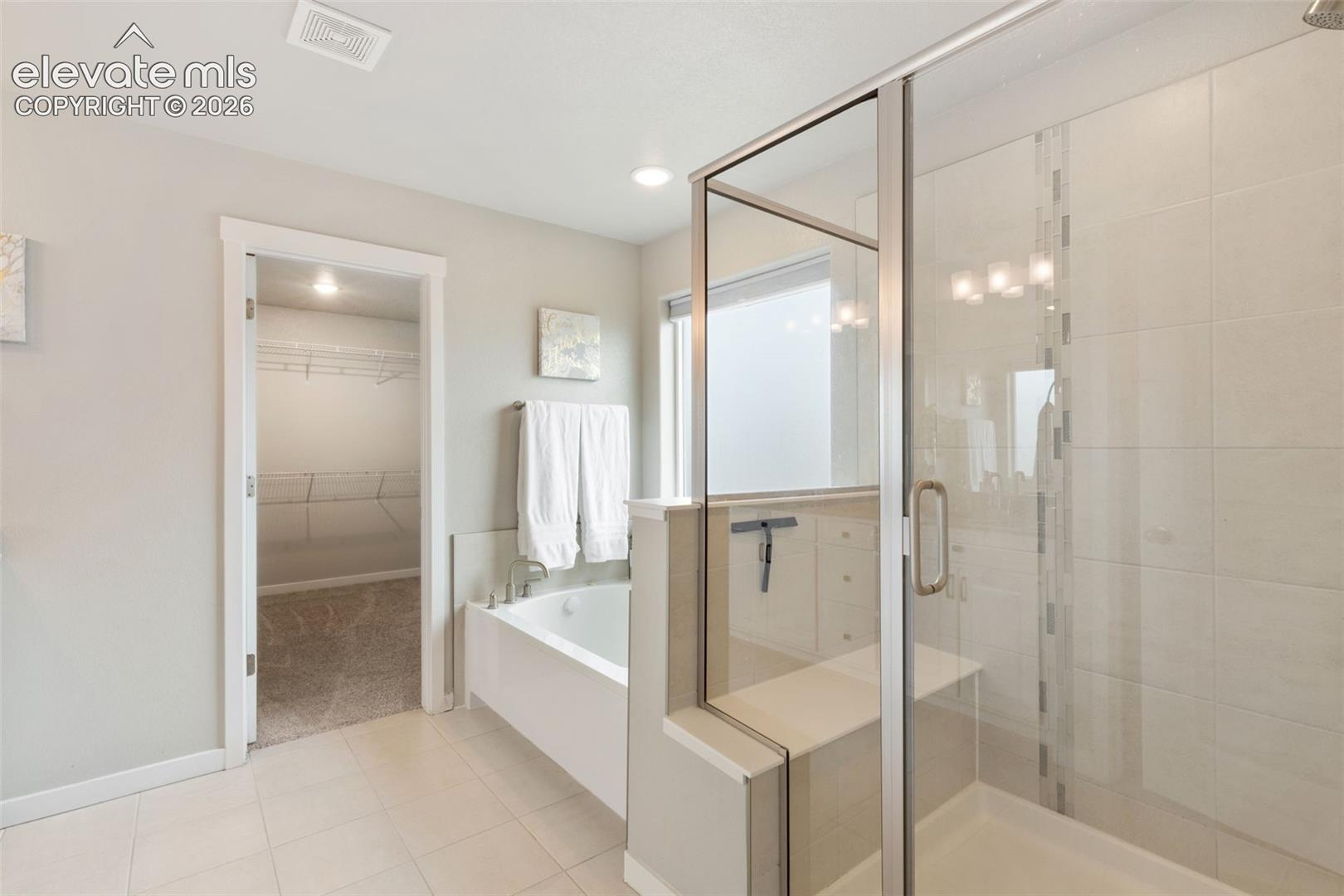 Bathroom featuring a glass-enclosed shower with an integrated bench, tile flooring, a separate soaking tub, and an opening to a walk-in closet with wire shelving