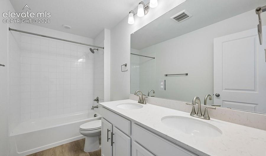 Dual-sink vanity with white cabinetry and stone-composite countertop, full-width wall mirror, three-light fixture, and tub-shower combination featuring white tile surround