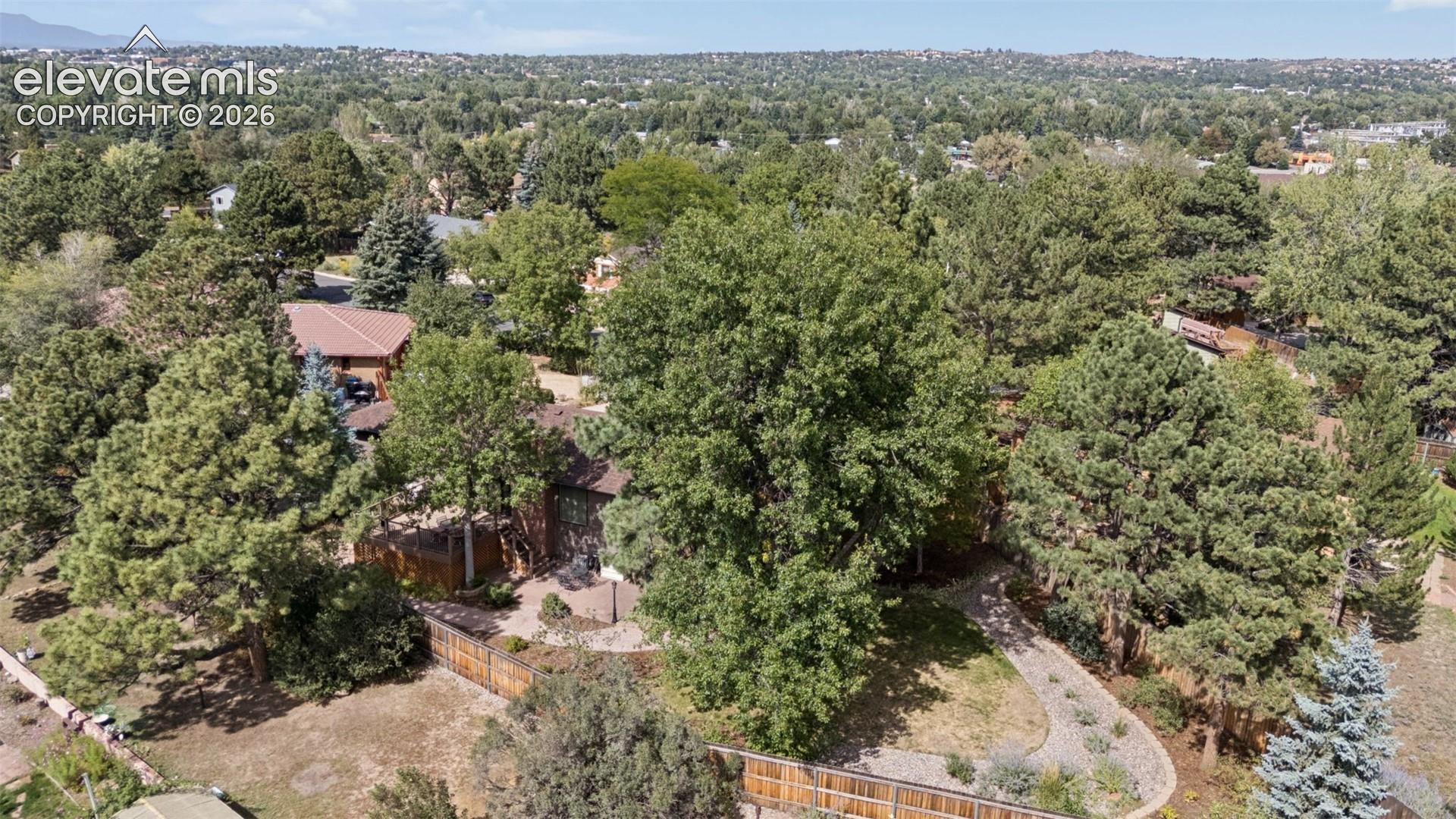 Aerial perspective featuring a multi-level residence surrounded by mature evergreen and deciduous trees, expansive landscaped grounds, stone-paved pathways, and wood perimeter fencing