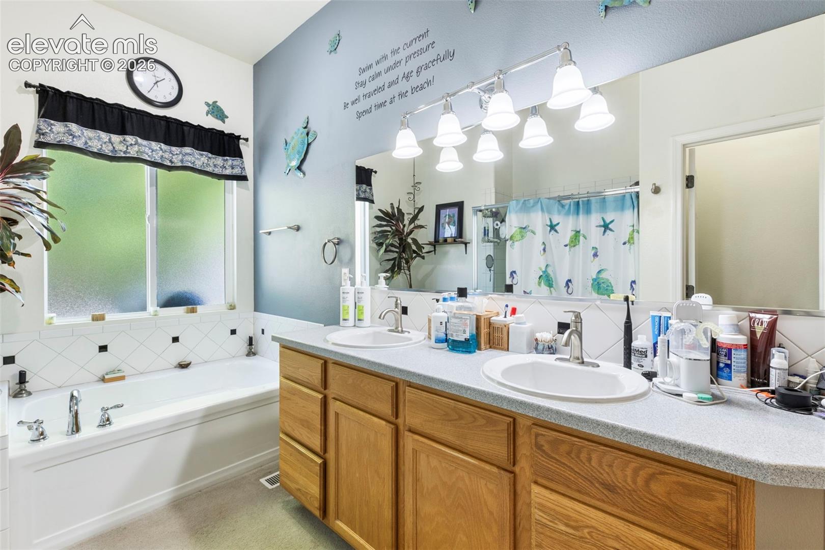 Double vanity with wood-finish cabinetry, built-in bathtub, window with privacy glass, and tiled wall accents