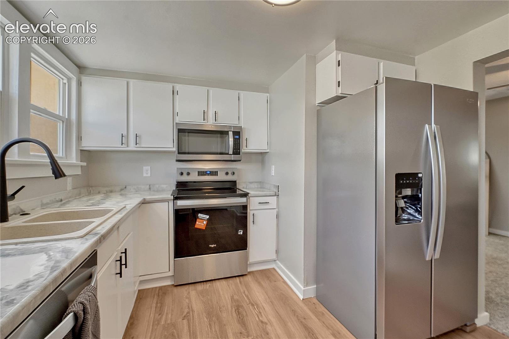 Kitchen featuring stainless steel appliances, wood-finish flooring, white cabinetry, a double basin sink with a matte black faucet, and light-toned countertops