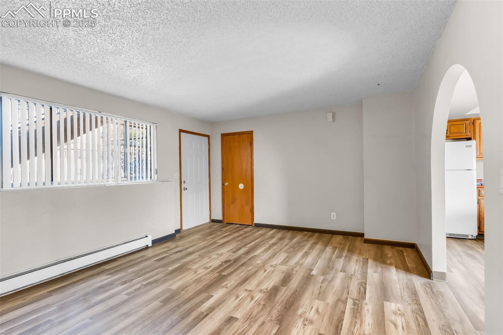 Empty room with light wood finished floors, arched walkways, a textured ceiling, and a baseboard radiator