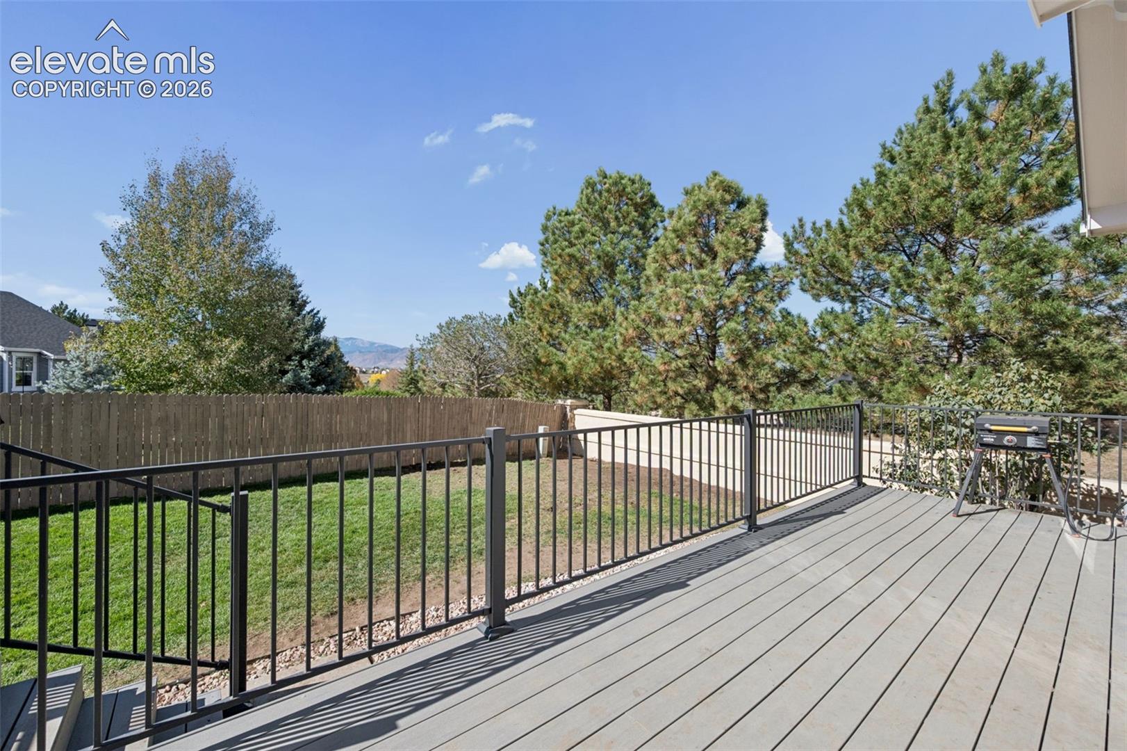 Elevated composite deck featuring a black metal railing, manicured lawn, perimeter fencing, and mature coniferous trees