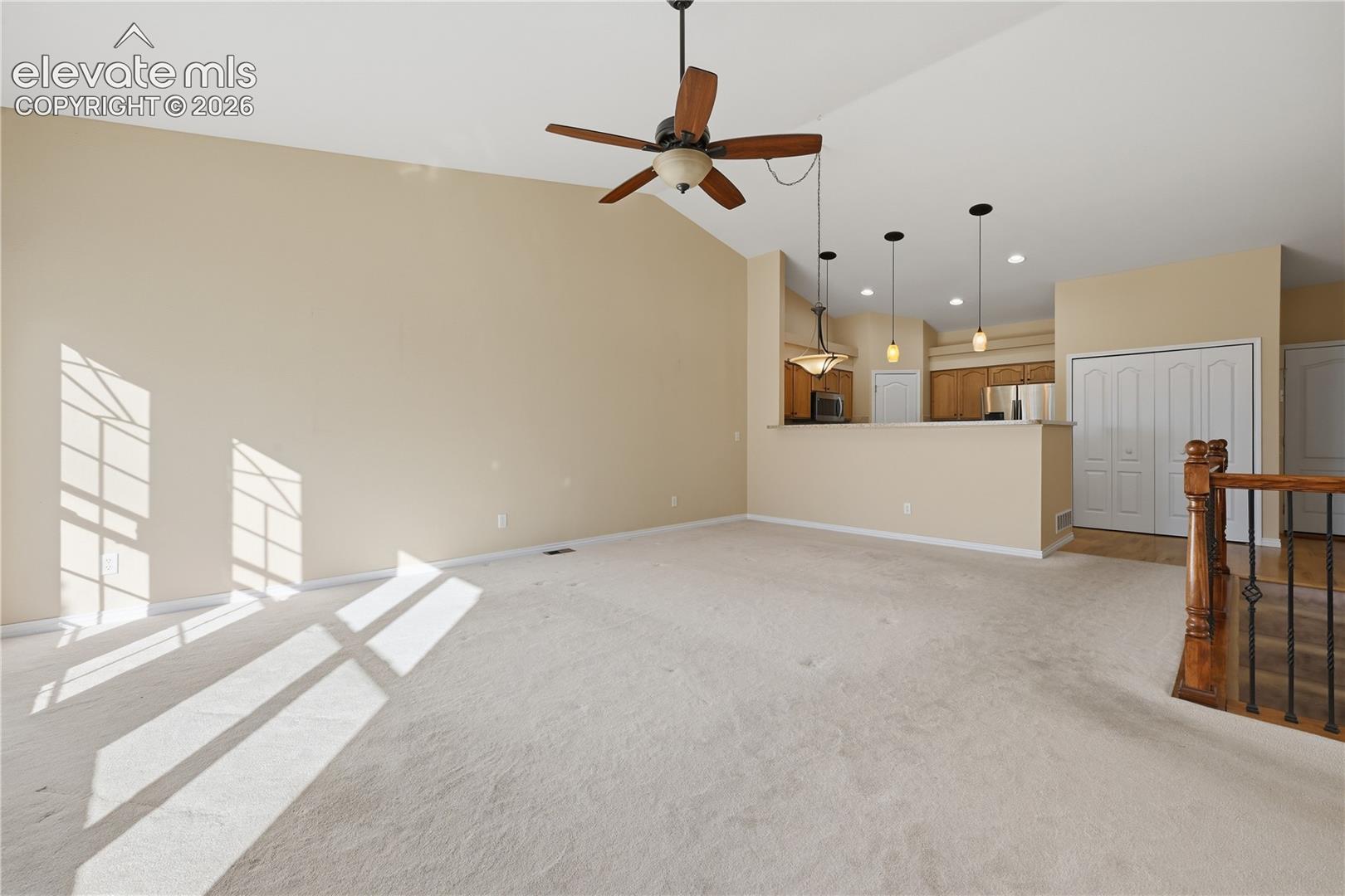 Vaulted ceiling featuring a central wood-blade ceiling fan, neutral wall color, overhead recessed lighting, three glass-pendant lights, and an interior staircase with a wood-finish handrail and metal balusters